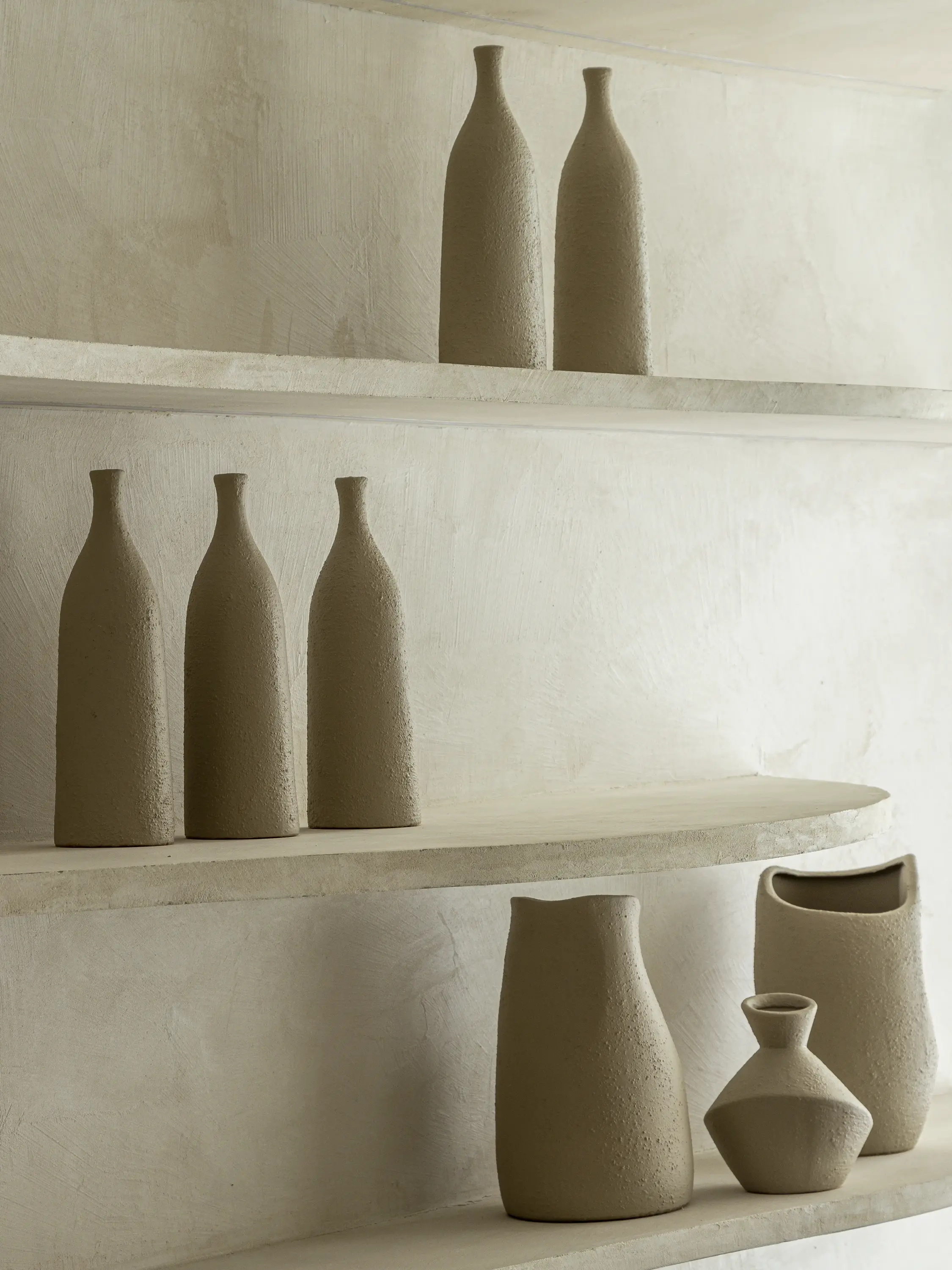 Prateleiras de concreto rústico exibindo cerâmicas artesanais em tons de bege. Na parte superior, garrafas alongadas de textura áspera; na inferior, vasos de formas orgânicas e minimalistas. O fundo apresenta uma parede com acabamento texturizado."