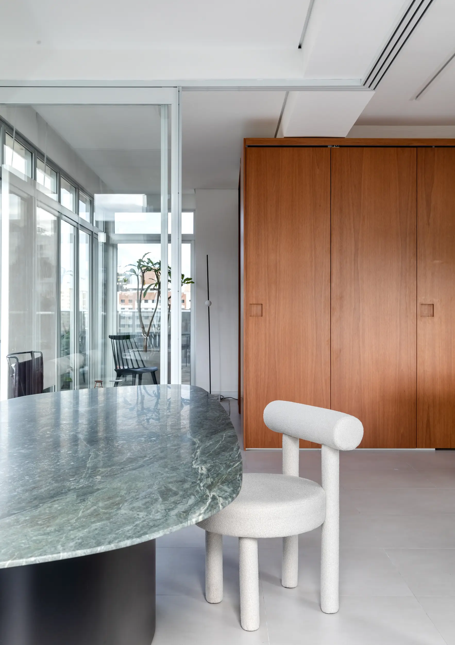 Ambiente contemporâneo com mesa de tampo em mármore verde de formato orgânico e base preta. Em primeiro plano, uma cadeira estofada em tecido claro com encosto cilíndrico. Ao fundo, armário de madeira de tom médio e divisória de vidro que revela uma varanda com cadeiras pretas e vegetação externa. Studio Guilherme Garcia.